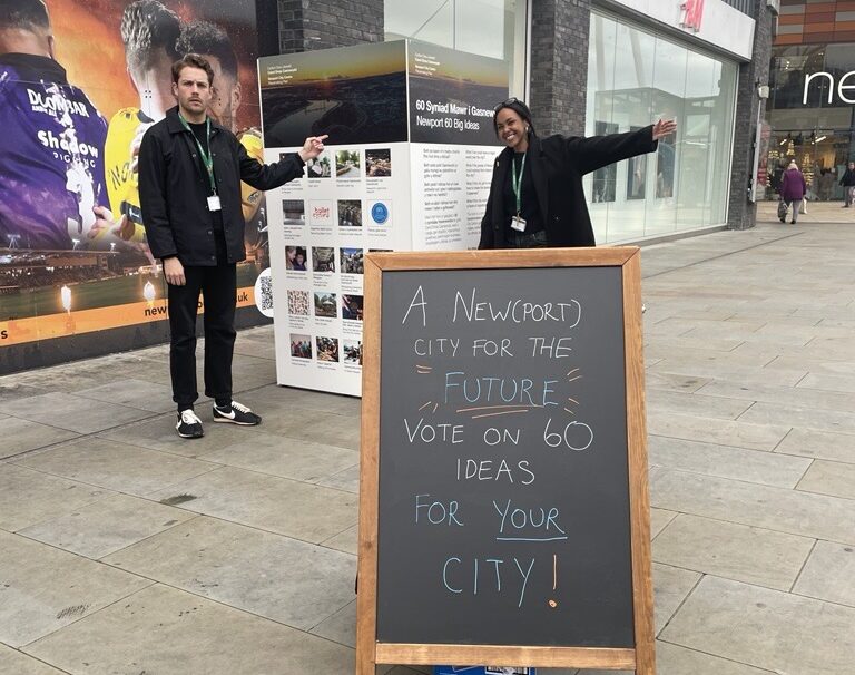 A black chalk board with text inviting people to engage with the community engagement team in the foreground and a man and woman in the background with open arms.