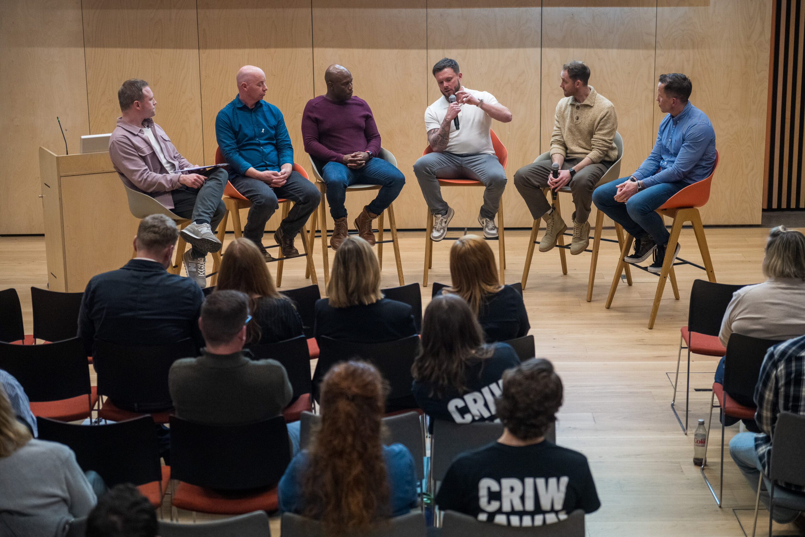 Six men sat on bar stools in a semi circle figure facing an audience 